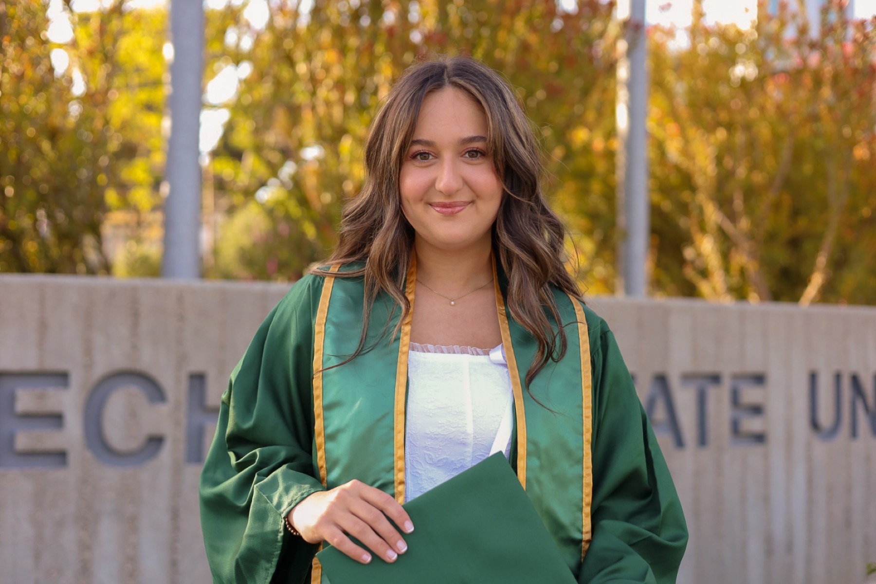 Graduate in a green stole in front of the Cal Poly university sign